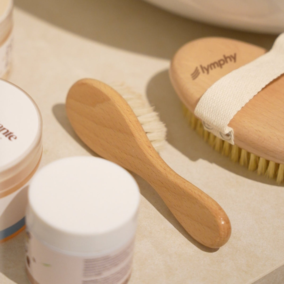 Flat-lay of the kit: face brush, body brush with cotton strap and balm jars on a beige surface
